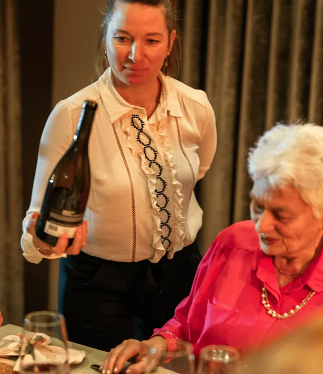 Een ouder koppel eet aan tafel, een ober ertussen staat klaar om wijn in te schenken.