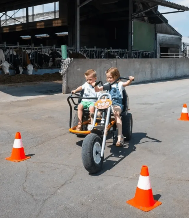 Jongen en meisje rijden op een go-cart en slalommen tussen kegels. In de achtergrond staan koeien in een stal.