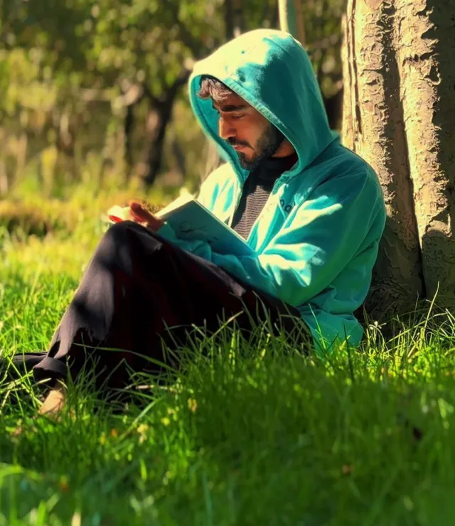 Man met kap op het hoofd zit in het gras tegen een boom en leest een boek