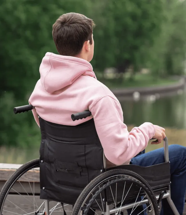 Jongen in een hoodie zit in een rolstoel en kijkt naar een vijver in een park